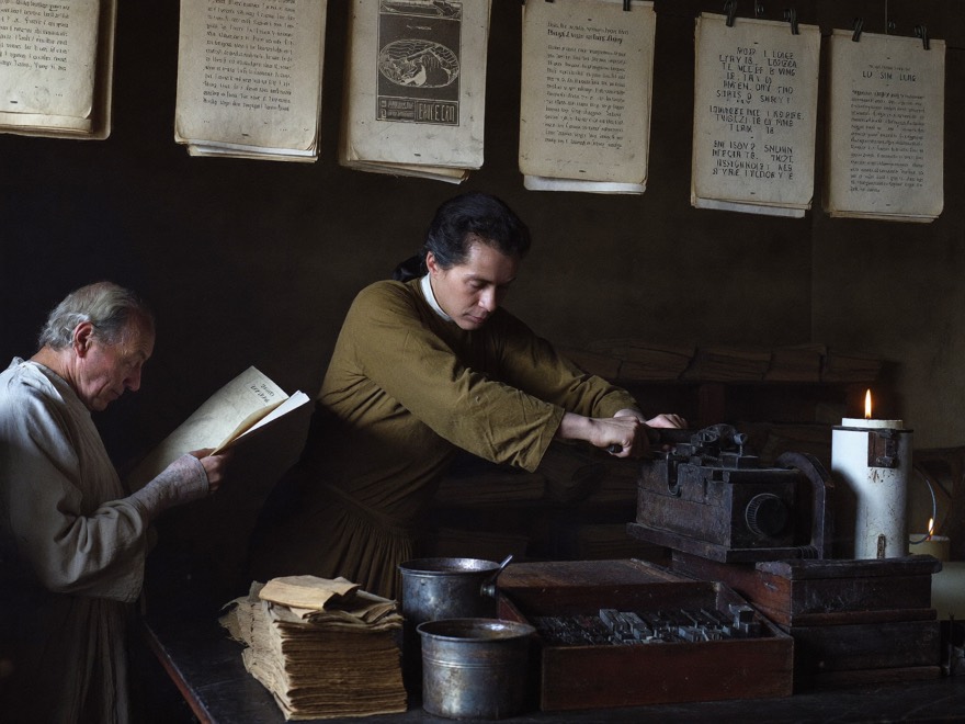 A printer works a wooden hand press by candlelight while a compositor reads a fresh proof, printed pages hanging above