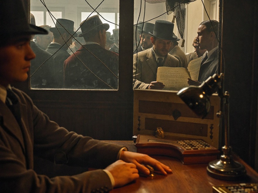 A telegraph operator at the brass key while a crowd of townspeople beyond the office glass reads the incoming message