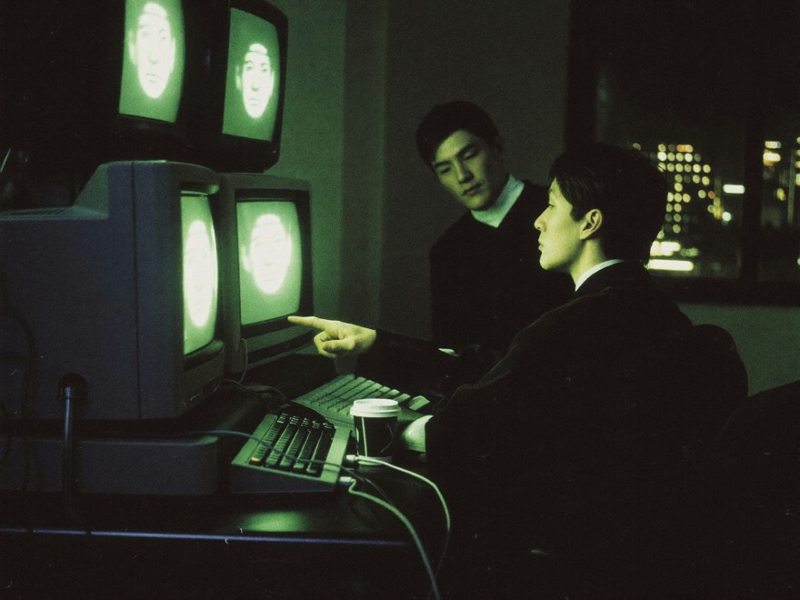 Two researchers in a dim 1991 computer room lit by the green glow of CRT terminals showing faces, city lights beyond the window